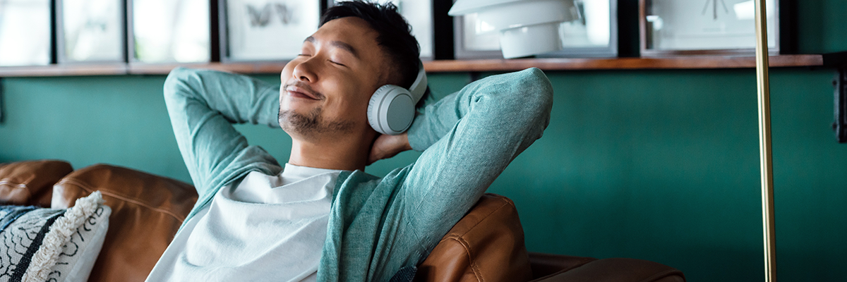 Young Asian man with hands behind head, relaxing on sofa and listening to music with headphones at home. Relaxed young man lying on sofa with music. Relaxing lifestyle, people and technology concept